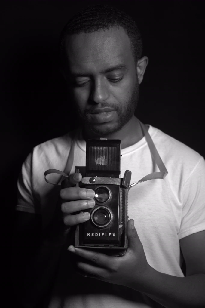 A man in a white T-shirt looks down at an old Ansco Rediflex camera held at his chest, shot in black and white against a dark backdrop.
